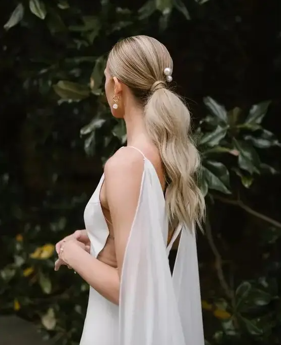a long low textural and wavy ponytail with a sleek top and pearls that accent the hairstyle is a cool idea for a modern bride