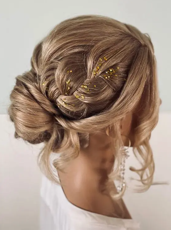 A lovely updo with a bun and large braids on top, a bump and face framing hair plus some gold glitter for an accent