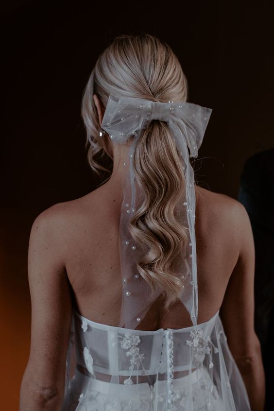 a low wavy ponytail with a sleek top and face-framing hair plus a sheer pearl bow on top is a cool idea to look trendy