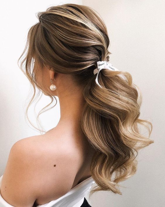 A pretty wavy low ponytail with a wavy top and some face framing hair plus a little bow is a chic and cool hairstyle for a modern bride