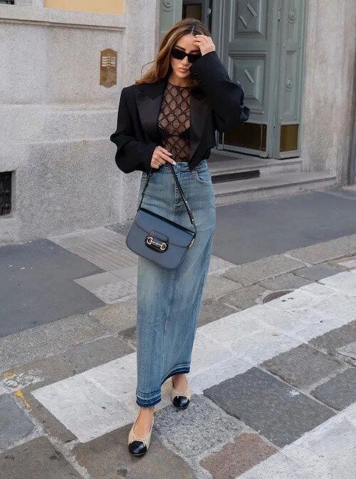 A sheer black lace top, a blue denim maxi skirt, a black cropped blazer, a bag and two tone shoes