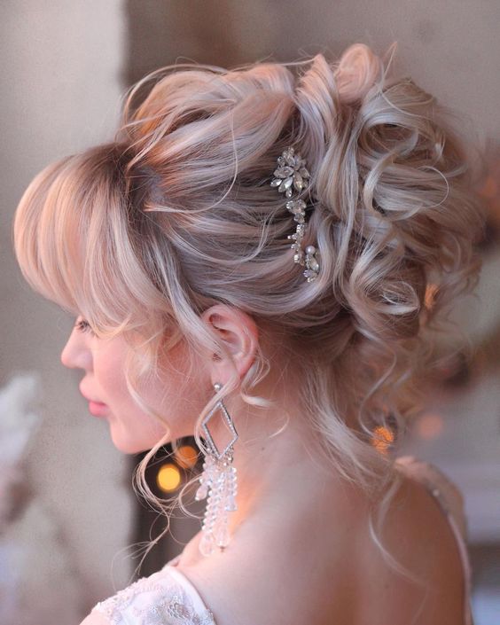 a sophisticated curly updo with a bump on top and layered bangs plus a rhinestone hairpiece is wow