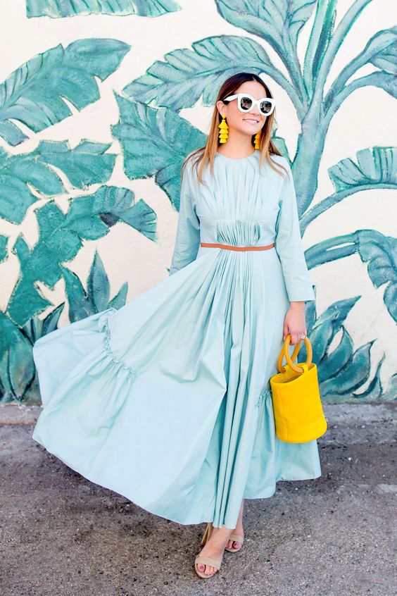 a spring wedding guest look with a blue pleated maxi dress, a belt, nude shoes, a bold yellow bucket bag and earrings
