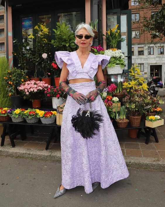 a whimsical spring wedding guest look with a lilac crop top with ruffles, a maxi skirt, a black feather bag and quirky sheer gloves