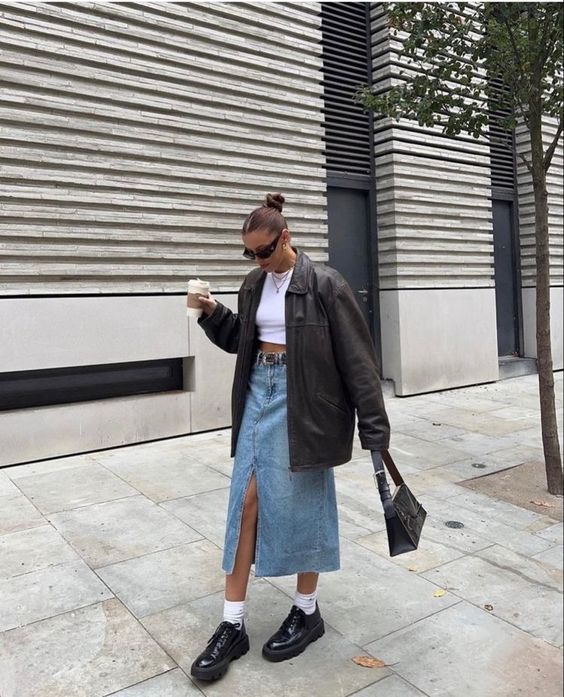 a white crop top, a blue denim midi skirt, a brown oversized leather jacket, black flat shoes and white socks and a small bag