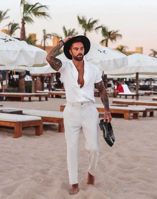 A beach wedding guest look with a white short sleeve shirt, neutral pants, black loafers and a black hat
