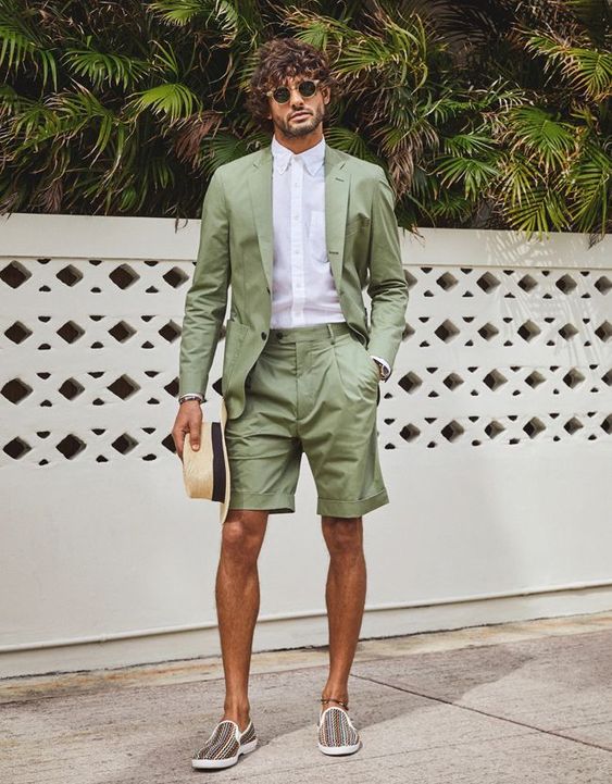 a cool beach wedding guest look with a green blazer and matching shorts, a white button down, printed loafers and a hat