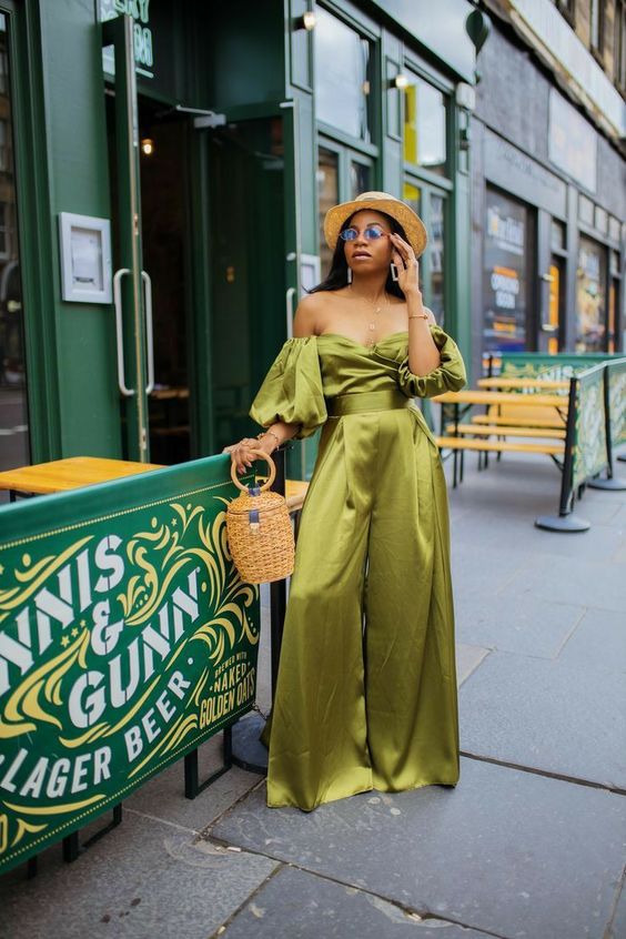 a satin green jumpsuit with puff sleeves, wideleg pants, a basket bag and a hat plus statement jewelry for a beach wedding