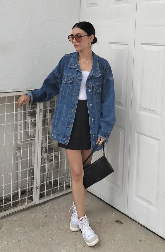 a spring look with a white top, a black mini, neutral sneakers, a blue oversized denim jacket, a black bag and chic jewelry