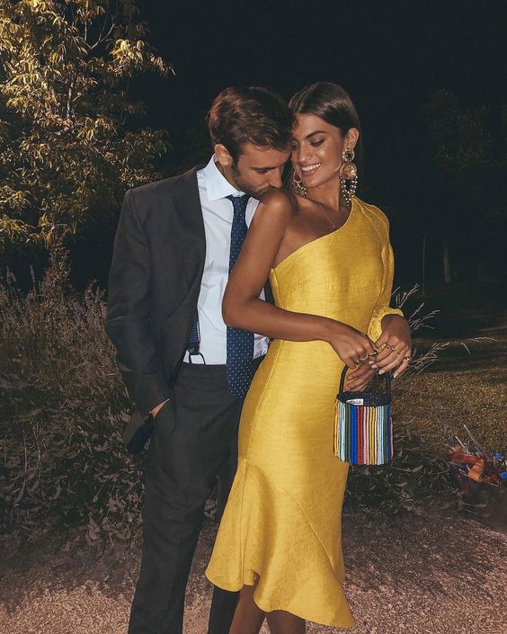 a yellow one shoulder midi dress with an asymmetrical skirt, a colorful bucket bag and statement earrings for a beach wedding